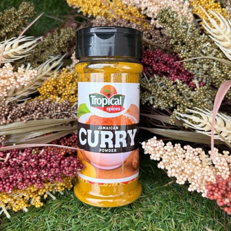 Tropical Spices Jamaican Curry Powder bottle on green grass surrounded by dried colorful plants.