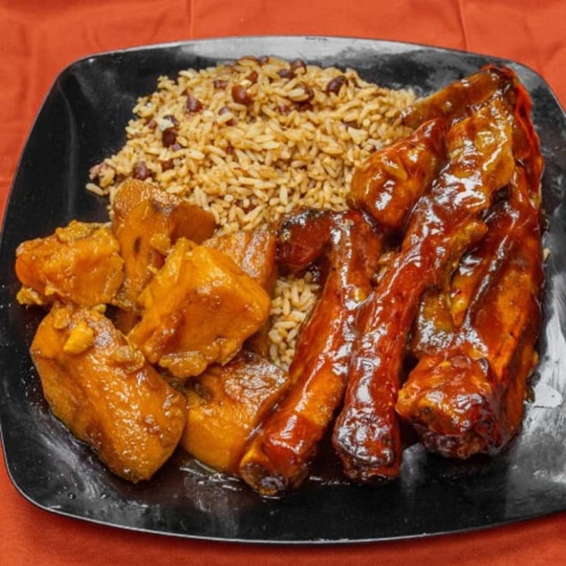 A black plate with ribs, rice and beans, and sweet potatoes on a red background.