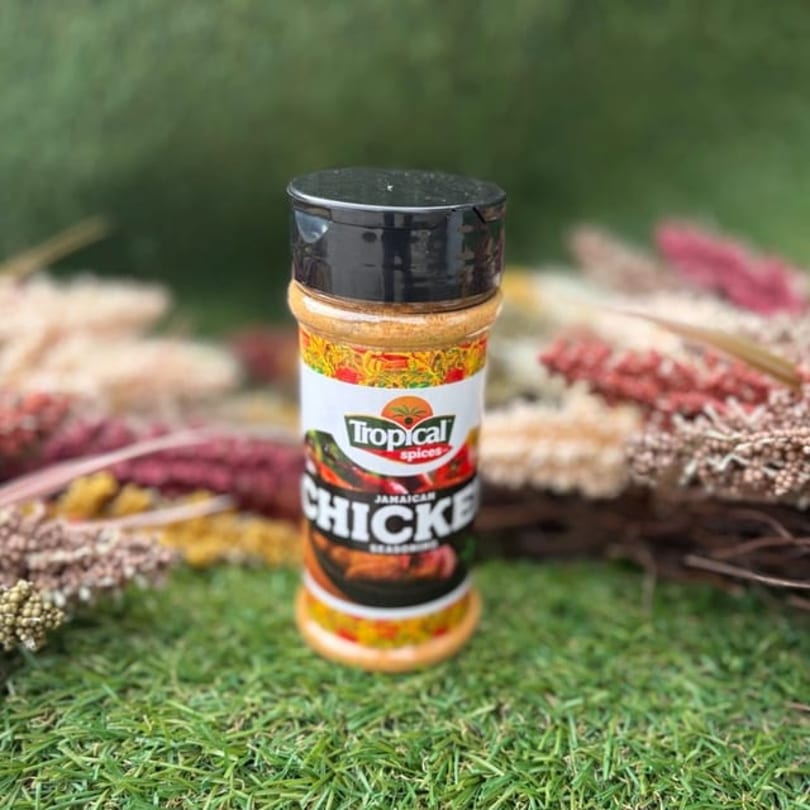 Tropical Spices Jamaican Chicken Seasoning bottle on green grass with blurred foliage.