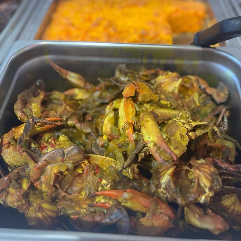 Close-up of a metal tray filled with Jamaican curry crabs, with another dish blurred in the background.