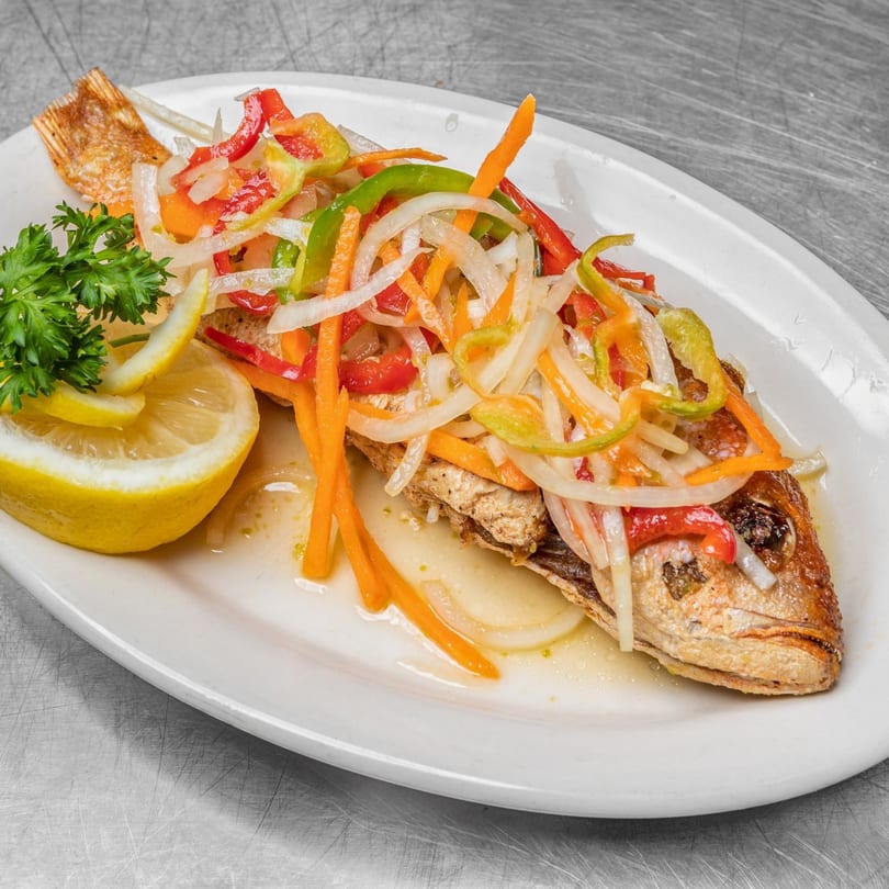 Whole fried fish topped with colorful pickled vegetables, lemon wedges, and parsley on a white plate.