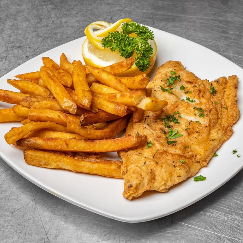 A white plate holds golden-brown fried fish and a pile of thick-cut french fries, garnished with lemon and parsley.