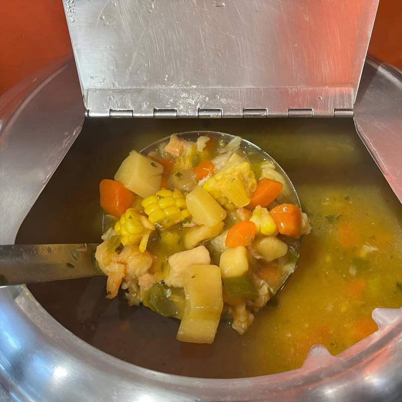 A ladle full of Jamaican seafood soup with corn, carrots, potatoes, and shrimp in a metal serving pot.