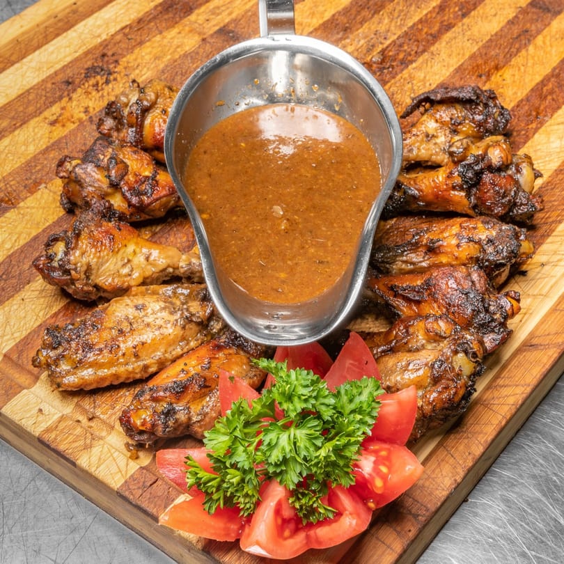 Grilled Jamaican jerk chicken wings with a side of sauce, garnished with tomatoes and parsley.