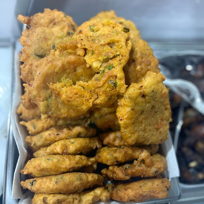A close-up of a large pile of golden-brown Jamaican fritters, some with visible herbs, in a serving tray.